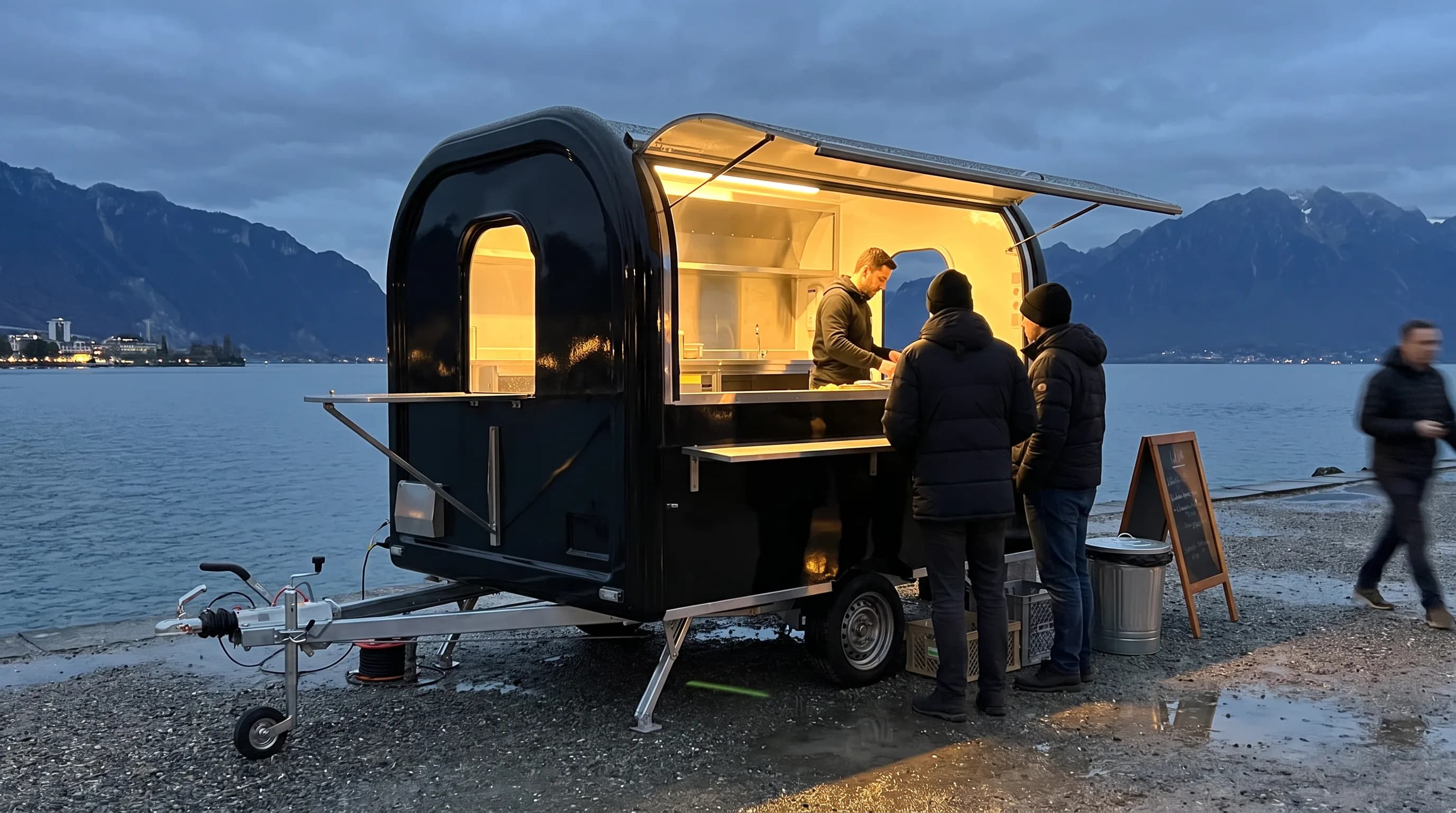 Black food truck trailer serving customers on the shore of Lake Geneva at dusk